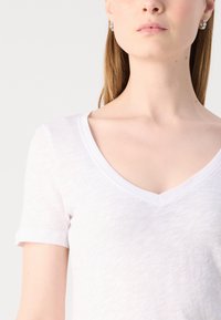 Fehér pamut póló V nyakkivágással és rövid ujjal; puha, texturált anyaggal rendelkezik, enyhén áttetsző megjelenésű.