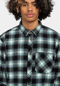 Grøn og sort ternet flannel skjorte med knap-til-design, brystlomme og tekstureret stof. Har rundet krave og brune knapper.