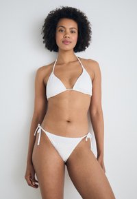 Hvit bikini-sett med en teksturert, heklet stil topp med trekantkopper og justerbare bånd, matchende lavt midje underbukser med sidesnøring.