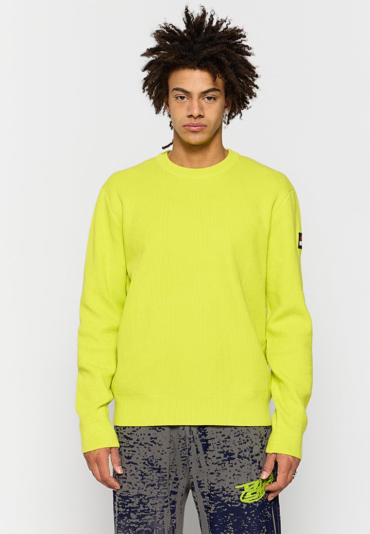 Maglione a coste giallo neon con scollo rotondo e maniche lunghe. Presenta una toppa con logo sulla manica sinistra. Indossato con pantaloni grigi a fantasia.