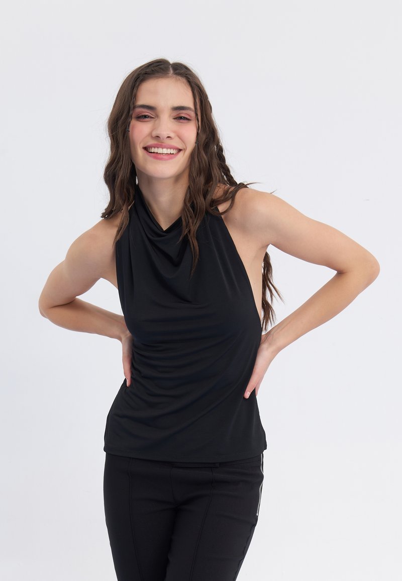 Crni halter top od mekog materijala s dizajnom s drapiranjem sprijeda. Ima bezrukav cut i glatku teksturu, uparen s crnim uskim hlačama.