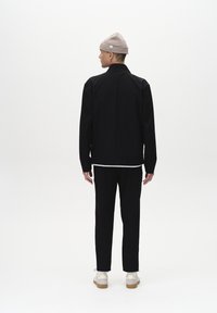Chaqueta negra con cuello alto, llevada sobre una camisa blanca, combinada con pantalones negros y zapatillas beige. Modelo con un gorro de punto beige claro.