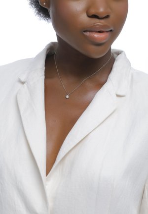 Gros plan d'une femme portant un petit pendentif en collier et un blazer blanc, avec un maquillage naturel et une couleur de lèvres neutre.