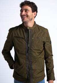 Giacca bomber verde oliva con zip frontale, due tasche sul petto e colletto rialzato; presenta un tessuto testurizzato e dettagli a contrasto.