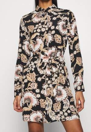 Robe-chemise noire à manches longues avec boutons, présentant un grand imprimé floral beige et blanc, et une ceinture nouée à la taille.
