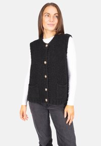 Schwarze, strukturierte Weste mit tiefem V-Ausschnitt, ausgestattet mit fünf goldenen Knöpfen und zwei Vordertaschen, getragen über einem weißen Langarmoberteil.