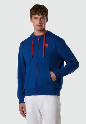 Giovane uomo con felpa con cappuccio blu con cerniera, cordini rossi e logo, mani in tasca, in piedi davanti a uno sfondo chiaro e semplice.