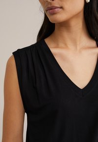 Kvinna i ärmlös svart V-ringad topp med plisserade detaljer nära axlarna mot en neutral bakgrund.