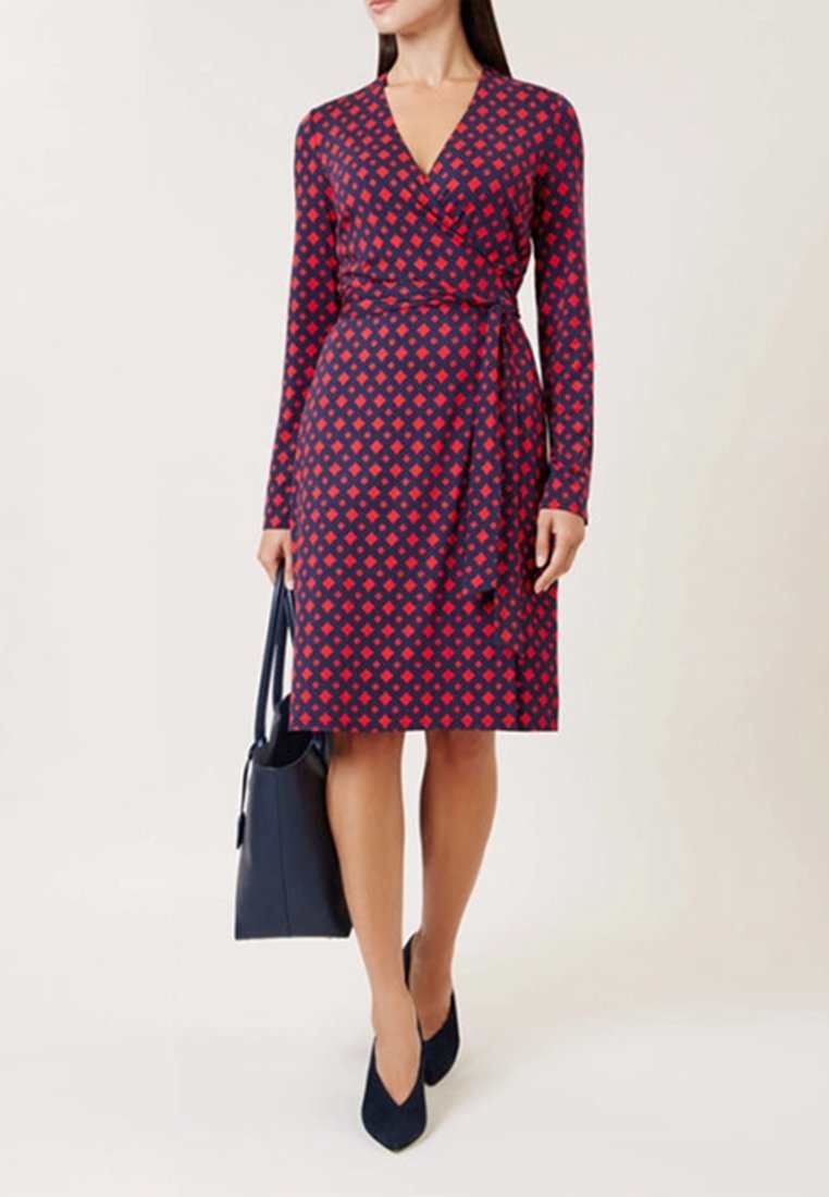 Femme portant une robe portefeuille bleu marine avec un motif losanges rouges, tenant un sac à main bleu marine et portant des talons pointus bleu marine.