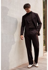 Mand i mørkebrun dragt og rullekrave, hvide sneakers, står på trædæk ved siden af lys betonvæg og kigger ned.