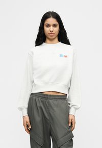 Γυναίκα με μακριά μαύρα μαλλιά που φοράει λευκό κοντό φούτερ με λογότυπο και γκρι παντελόνι cargo, στέκεται μπροστά σε απλό φωτεινό φόντο.