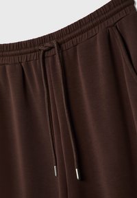 Bruna joggerbyxor i mjuk tyg, med elastisk midja, snöre med metalländar och sidofickor. Slät textur.