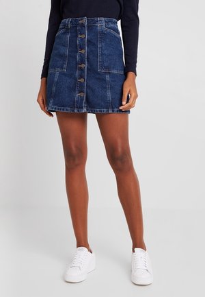 Frau trägt ein dunkelblaues Langarmshirt, Mini-Jeansrock mit Knopfleiste und Taschen sowie weiße Turnschuhe, steht vor einem einfarbigen Hintergrund.