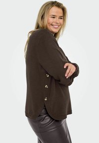 Femme souriante avec de longs cheveux blonds, portant un pull en tricot marron à boutons et une jupe en cuir noir, les bras croisés, tournée légèrement de côté.