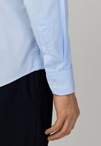 Męskie ramię w jasnoniebieskiej koszuli z mankietem na guziki, spoczywające obok czarnych spodni na neutralnym tle.