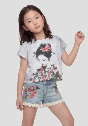 Niña joven con cabello ondulado que lleva una camiseta gris con una ilustración de una geisha y pantalones cortos de mezclilla con bordado floral colorido y borde de encaje.