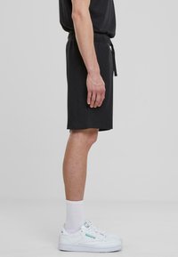 Shorts noirs en coton avec une taille élastique et un cordon de serrage réglable ; texture lisse ; associés à des baskets blanches et des chaussettes côtelées.