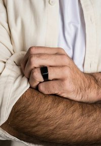 Schwarzer Ring mit rechteckiger Form an einer Hand, die auf einem Unterarm ruht. Der Träger hat helles Haar und trägt ein hellbeiges Hemd.
