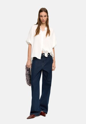 Biała, oversize koszula na guziki, zestawiona z ciemnoniebieskimi jeansami o szerokich nogawkach. Modelka trzyma szarą kopertówkę z futra. Brązowe buty z ostrym noskiem.