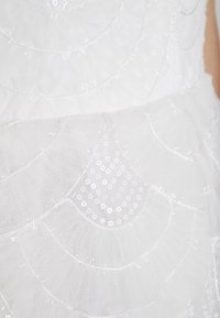 Robe blanche en tulle superposé, présentant des motifs courbés et des sequins. Surface texturée avec des accents brillants et des détails de couture délicats.
