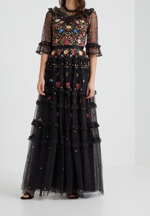 Kobieta ubrana w czarną, przezroczystą suknię maxi z kolorowym kwiatowym haftem, falbanami i rękawami do łokcia, stojąca na jednolitym tle.