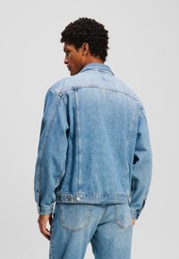 Ljusblå denimjacka med klassisk krage, knappstängning och två sidofickor, med sömnadsdetaljer och logotyp på baksidan.