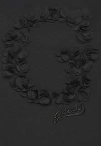 Camiseta negra con una textura floral en relieve que forma una figura en forma de "G". Presenta el logo bordado "Guess" con una tipografía elegante en la parte inferior.