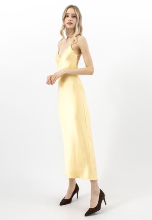 Kobieta ubrana w żółtą, satynową sukienkę midi na ramiączkach typu spaghetti i czarne szpilki, stojąca bokiem na białym tle.