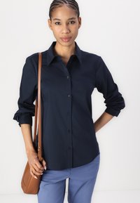 Navy blue button-up shirt with a collared neck, rolled sleeves, and a smooth texture; paired with light blue pants and a brown crossbody bag.