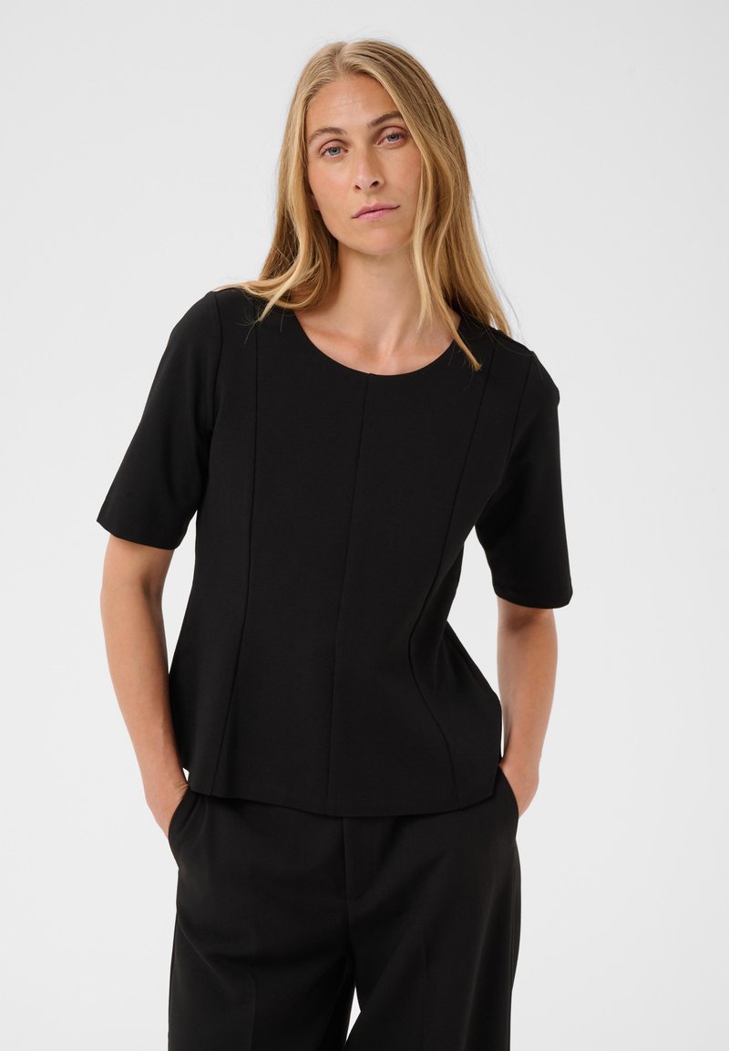 Femme aux longs cheveux blonds portant un haut noir ajusté à manches courtes et un pantalon noir, les mains dans les poches, sur un fond uni.