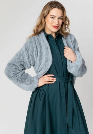 Kobieta o długich blond włosach, ubrana w turkusową sukienkę z paskiem i jasnoniebieski, gruby sweter na drutach, pozująca z jedną ręką na biodrze, a drugą trzymającą sweter.