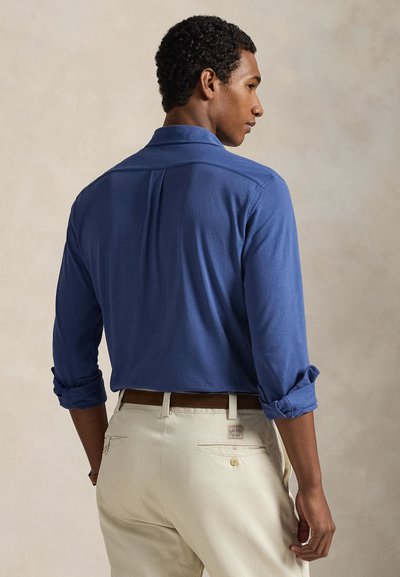 Βασιλικό μπλε πουκάμισο με κουμπιά, με μανίκια γυρισμένα, απαλής υφής και με πιέτα στην πλάτη, συνδυασμένο με μπεζ παντελόνι και καφέ ζώνη.