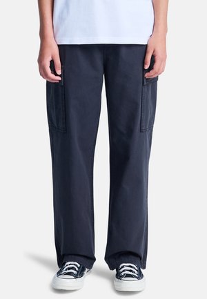 Personne portant un pantalon cargo noir ample, des baskets noires et blanches et une chemise blanche, debout devant un fond blanc.