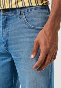 Jeans in denim azzurro chiaro presentano un design a gamba dritta, cinque tasche e cuciture a contrasto, con una mano appoggiata sulla coscia.