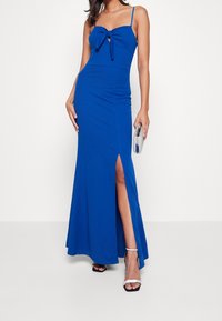 Blå golvlång klänning med en åtsittande livdel, en rosett detalj vid halsringningen och en slits på sidan. Kompletterad med silverfärgade klacksandaler och en clutch.