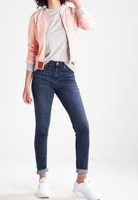 Žena v světlém růžovém bomberu, šedém tričku, tmavě modrých skinny jeans s ohrnutými nohavicemi a bílých teniskách, stojící na bílém pozadí.