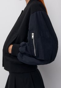 Veste bomber noire avec manches bleu marine bouffantes, poignets côtelés et poche zippée sur le bras gauche. Tissu lisse, coupe courte décontractée.