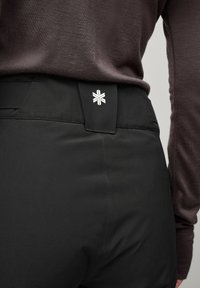 Black pants with a smooth texture, featuring a tailored fit and a discreet logo patched on the waistband. Close-up view of the back.