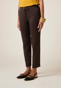 Pantalons marron sur mesure à coupe ajustée et ourlets à revers, avec un détail de bouton doré. Associés à des ballerines noires à bout pointu et des bracelets colorés.