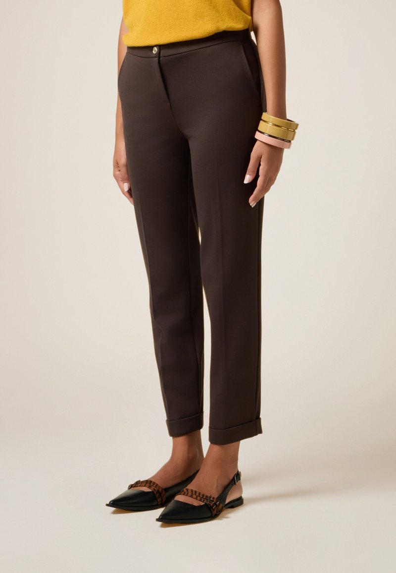 Pantalons marron sur mesure à coupe ajustée et ourlets à revers, avec un détail de bouton doré. Associés à des ballerines noires à bout pointu et des bracelets colorés.