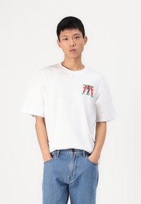 Tricou alb din bumbac, cu o croială lejeră, având un design grafic colorat pe partea stângă a pieptului, purtat cu blugi bleu.
