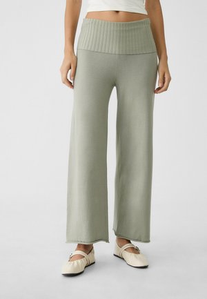 Osoba nosząca jasnozielone, szerokie spodnie z wysokim stanem oraz kremowe baleriny Mary Jane, stojąca na jasnym, jednolitym tle.