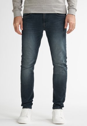 Man draagt donkerblauwe slim-fit jeans, witte sneakers en een lichtgrijze sweatshirt, staand tegen een effen witte achtergrond.