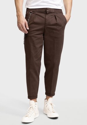 Mann trägt dunkelbraune, zulaufende Hose mit einer silbernen Kette auf der linken Seite, weiße High-Top-Sneakers und ein teilweise sichtbares weißes Hemd.