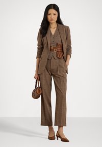 Ruskea tweed-puku, jossa on istuva bleiseri ja leveälahkeiset housut. Teksturoitu liivi napilla, jota korostaa ruskea vyö. Vaaleanruskea käsilaukku ja korot täydentävät asusteen.