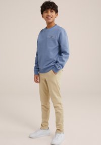 Világoskék pulóver kerek nyakkal és zsebfedikével, b�ézs, slim-fit nadrággal és fehér sportcipővel párosítva semleges háttér előtt.