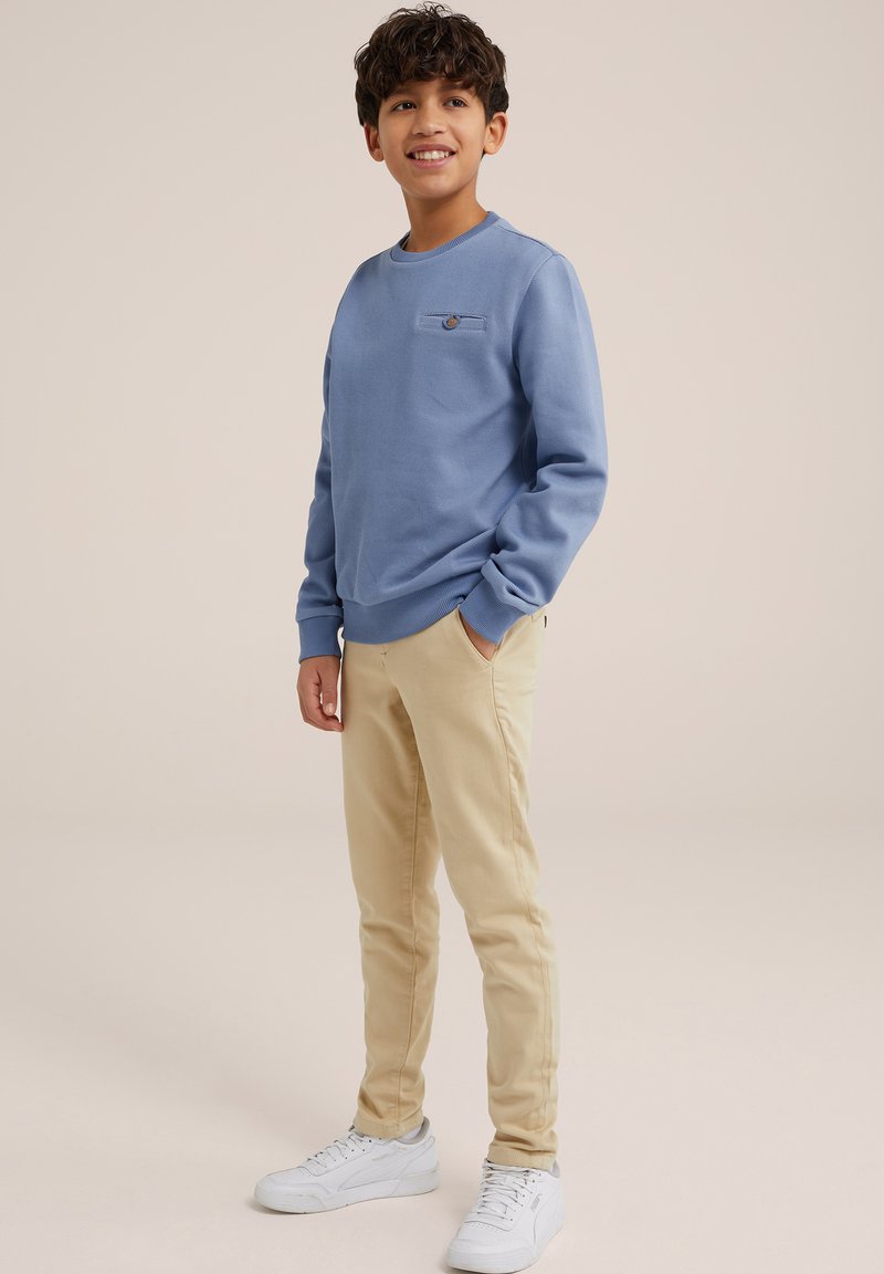 Világoskék pulóver kerek nyakkal és zsebfedikével, bézs, slim-fit nadrággal és fehér sportcipővel párosítva semleges háttér előtt.