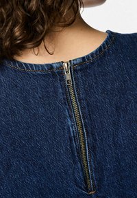 Jeansplagg med rund halsringning, utrustad med en synlig metalldragkedja på ryggen, framhävd med kontrasterande orange sömmar.