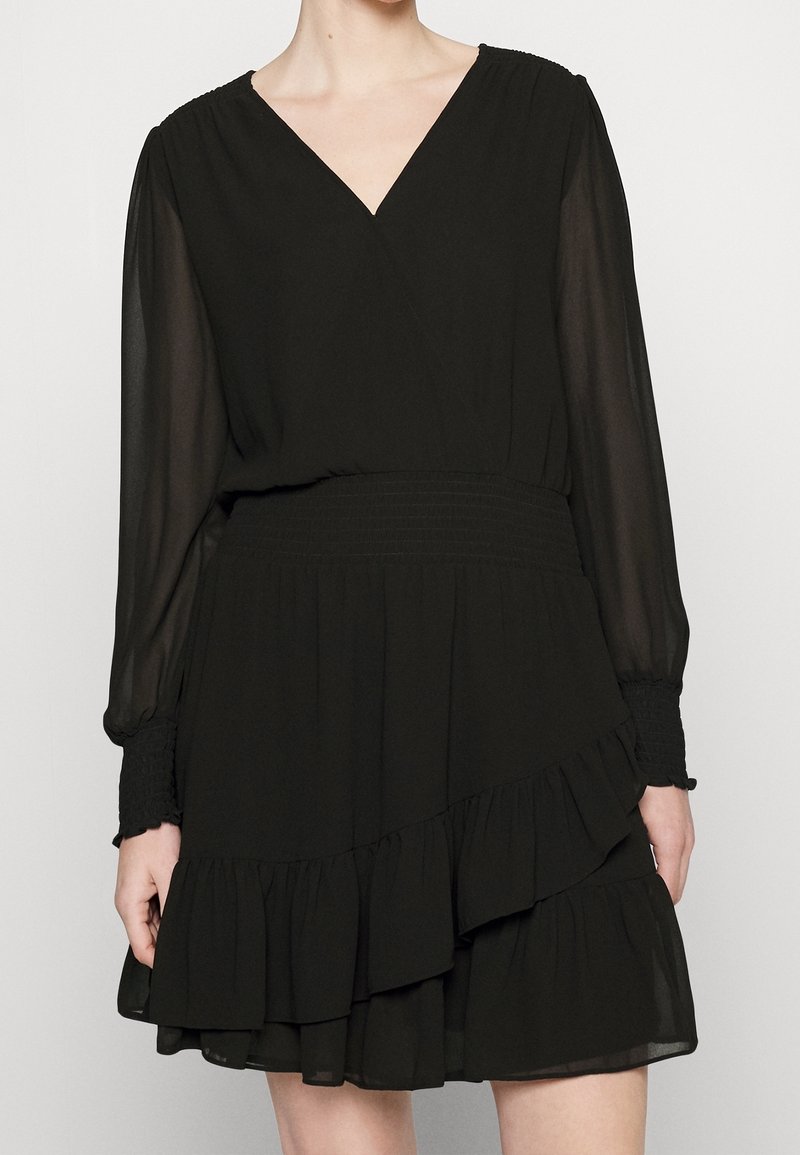 Femme portant une robe noire à manches longues avec un col en V, une taille froncée et une jupe à volants superposés, présentée du cou jusqu'à mi-cuisse.