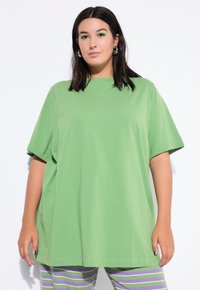 Kobieta z ciemnymi włosami, ubrana w oversize'owy zielony t-shirt, nosząca kolczyki w kształcie kół oraz zielony makijaż oczu, stoi na tle białej ściany, w pasiaste spodnie w kolorze purpurowym i zielonym.